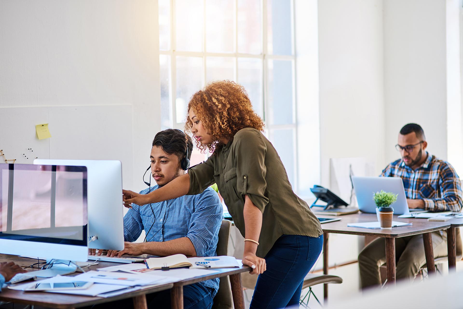 Three people in an office, with a woman helping a man with something on the computer